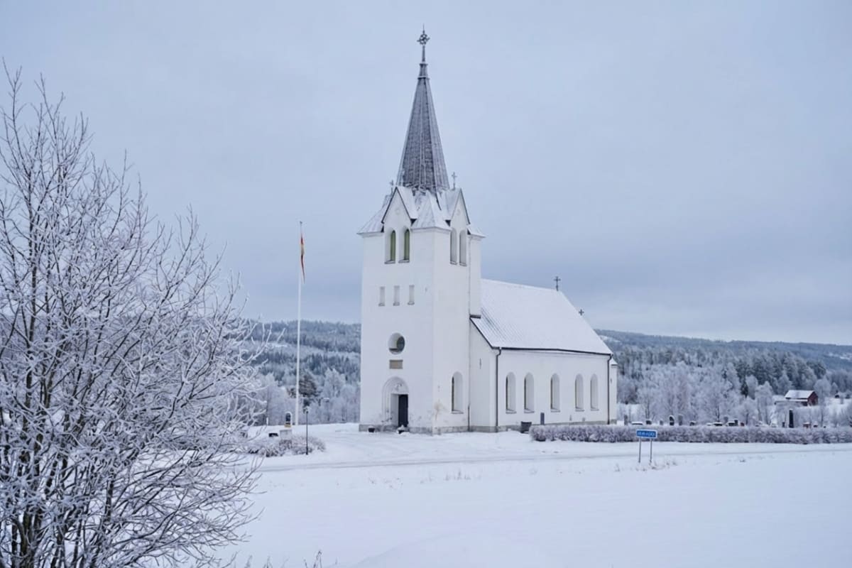 En vit kyrka med en spetsig kyrktorn, täckt med snö, omgiven av ett vinterlandskap. Träd täckta med snö och rimfrost syns i förgrunden och bakgrunden. En flaggstång står bredvid kyrkan.