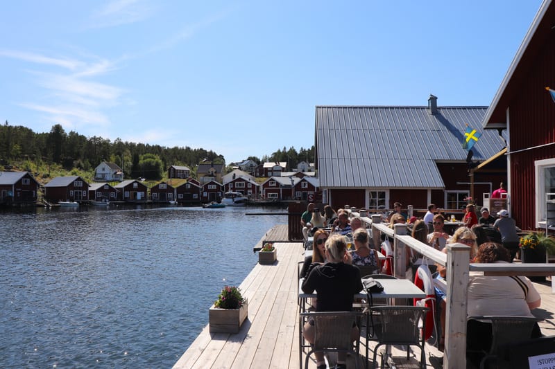En utomhusbild som visar en trädäckad restaurang eller servering vid en vattenkant fylld med gäster. Mittemot över vattnet syns röda sjöstugor och hus. Två svenska flaggor är synliga på en byggnad intill restaurangens uteservering. Det är en solig da