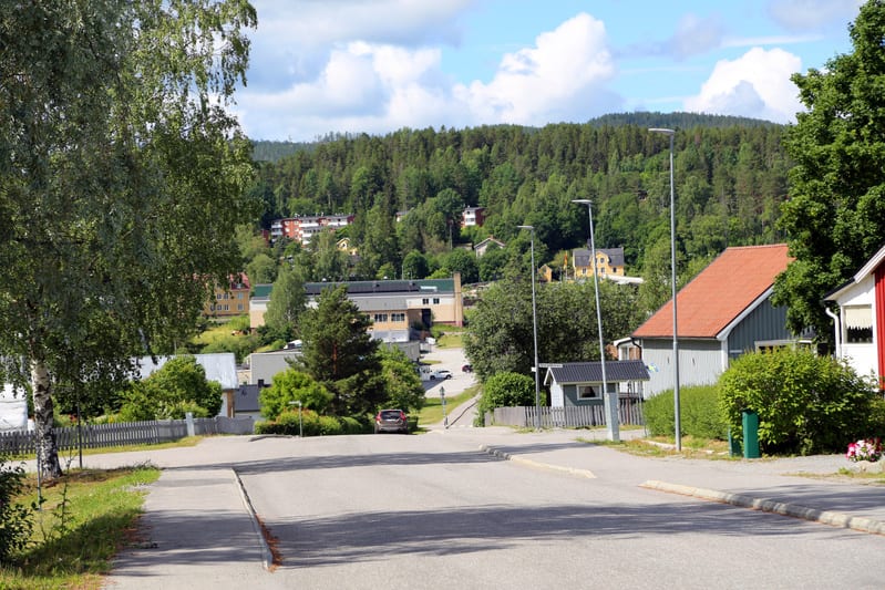 En gata leder nedåt mot ett blandat bostads- och industriområde, omgivet av grönskande träd och kullar med bebyggelse i bakgrunden under en blå himmel med moln. En vit bil står parkerad på en uppfart.