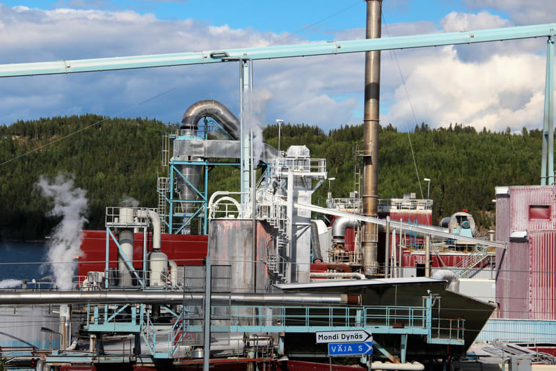 En industriell anläggning med omfattande rörsystem, tankar, skorstenar och olika byggnadsstrukturer. Vit ånga stiger upp från flera punkter. I bakgrunden syns skogklädda kullar och en del av ett vattendrag. En blå vägskylt visar riktning. Himlen är d