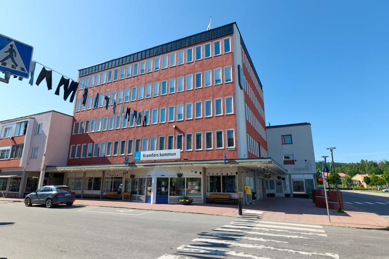 En röd byggnad med många fönster dominerar bilden under en klarblå himmel. Framför byggnaden finns en skylt med texten "Kramfors kommun" och flera kläder hänger på en lina. En personbil är parkerad till vänster och ett övergångsställe syns i förgrund