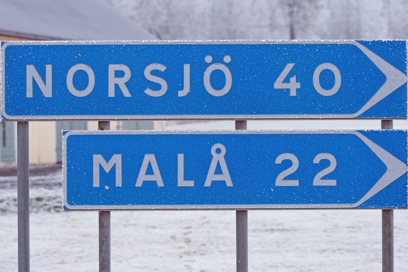 En vägskylt med blå bakgrund och vit text som visar riktningen till Norsjö 40 km och Malå 22 km. Skylten är täckt av frost, och i bakgrunden syns snö på marken samt delar av en byggnad med en ljus färg.