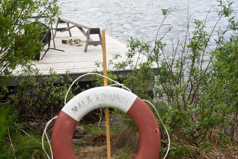 En livboj med texten "MALÅ KOMMUN" hänger på en träpåle i förgrunden, omgiven av gräs och buskar. I bakgrunden syns en brygga med en bänk och en grill, samt vatten som reflekterar ljus.
