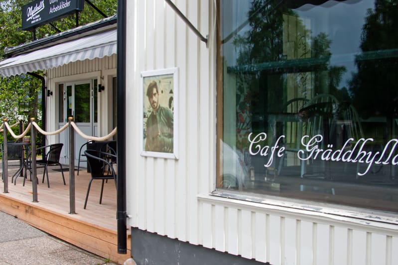 En bild som visar delar av ett vitt trähus med en uteservering på en träveranda. Byggnaden har en markis och skyltar. Ett fönster på byggnaden reflekterar omgivningen och en logotyp, 'Cafe Gräddhyllan', är synlig på fönstret.