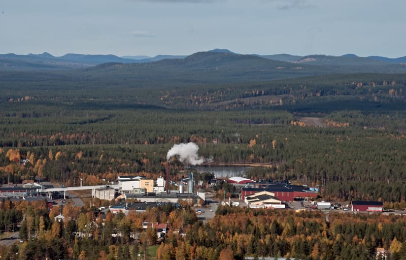 En vidvinkelbild tagen från ovan som visar en bebyggelse med industrikomplex, hus och vägar omgivna av omfattande skogsområden. I bakgrunden syns skogsklädda berg. En vattenyta och rök syns vid industriområdet, och lövträd i bilden har höstfärger.