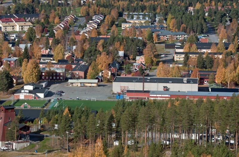 En vidvinkelbild från ovan som visar en del av ett samhälle under hösten, med bostadshus, en fotbollsplan med konstgräs, en större industribyggnad och skog med lövträd i höstfärger samt barrträd.