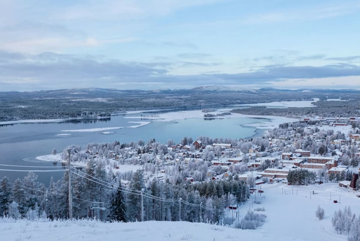 En vidvinkelbild från ovan som visar ett vinterlandskap med ett delvis isbelagt vattendrag omgärdat av snötäckta träd och byggnader. I förgrunden syns snöklädda trädtoppar och en del av något som ser ut som en skidlift.