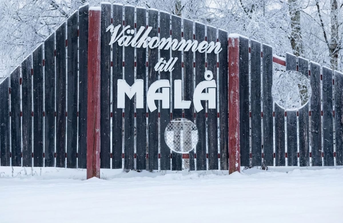 En svart träskylt med texten 'Välkommen till MALÅ' i vit färg, delvis täckt av snö. Skylten har även en transparent, rund detalj med ett nätmönster. Marken framför skylten är täckt av ett tjockt lager snö och i bakgrunden syns snötäckta träd.