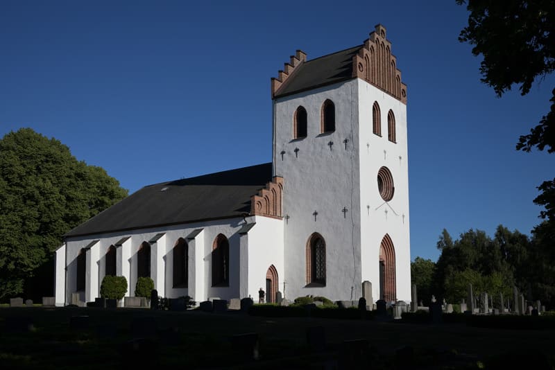 En vit kyrka med tegeltak och en framträdande klocktorn avtecknar sig mot en klarblå himmel. Kyrkan har flera fönster och en del av fasaden är i skugga. Träd med grönt lövverk omger kyrkan, och i förgrunden syns skuggiga gravstenar på en gräsmatta. E