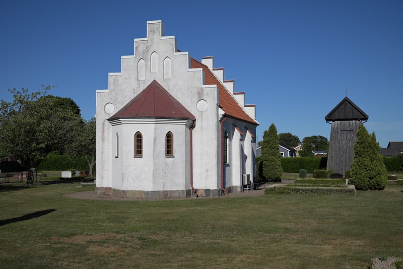 En vit kyrka med rött tegeltak och en takgavel i trappstegsform syns på en grön gräsmatta under en klarblå himmel. Till höger om kyrkan står en träkyrkklockstapel omgiven av gröna buskar och träd. I bakgrunden syns hustak och träd.