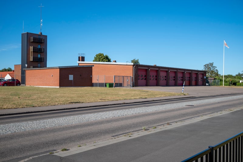 En röd tegelbyggnad med en ansluten mörkbrun byggnad med fönster och balkonger, mot en klarblå himmel. Stora röda garagedörrar med små fönsterrutor syns. Ett staket och en del av en bil syns också, samt en flaggstång med hissade flagga. En väg och gr