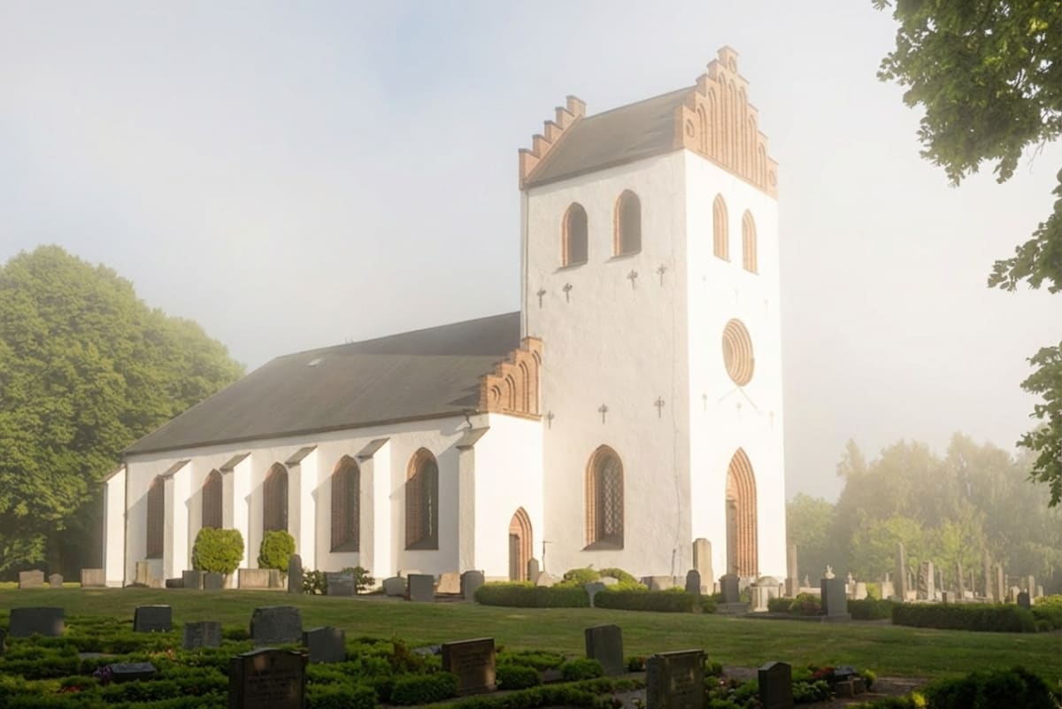 En vit kyrka med tegeldetaljer och ett mörkt tak står omgiven av grönska och en kyrkogård, delvis insvept i dimma. Gröna träd syns på båda sidor av bilden och himlen är ljus och disig. Gravstenar kan ses i förgrunden.