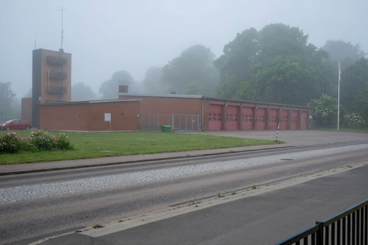 En stor röd tegelbyggnad med flera röda portar syns i dimma. Byggnaden har en hög fönsterlös del med en metallstruktur. En röd personbil syns parkerad framför byggnaden. Ett träd är synligt bakom byggnaden och en flaggstång utan flagga står vid sidan