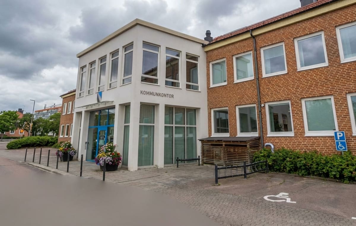 En bild som visar framsidan av en byggnad med texten "KOMMUNKONTOR" vid entrén. Byggnaden består dels av en ljus del med stora fönster och en röd tegeldel. Utanför finns blommor i krukor, en trottoar och en parkeringsplats med handikappsymbol.