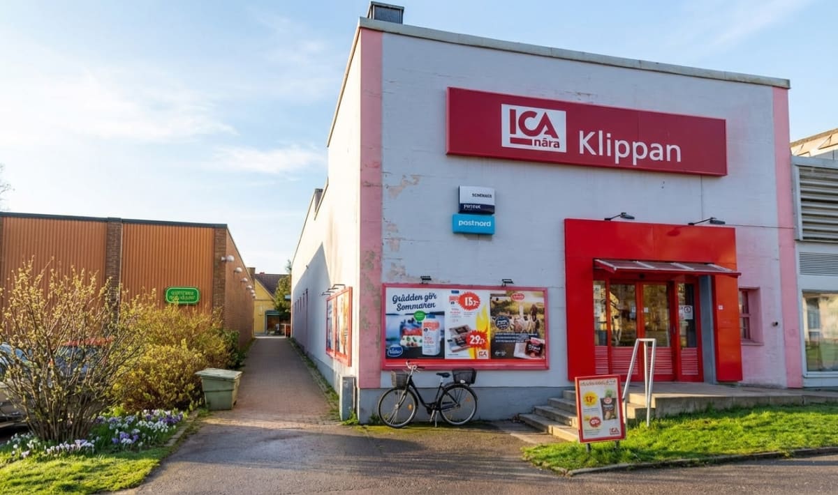 En bild som visar en ICA Nära Klippan-butik med röd logotyp och entré. En cykel är parkerad vid fasaden med reklam som annonserar "Grädden gör Sommaren". Blommande buskar och blommor syns i förgrunden, och en gångväg leder iväg längs en byggnad till