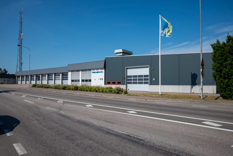 En modern byggnad med flera garageportar, en kommunikationsmast och en flaggstång med en flagga syns under bar himmel. En asfaltväg med vita markeringar löper framför byggnaden, och en grön buskrad kantar byggnaden.