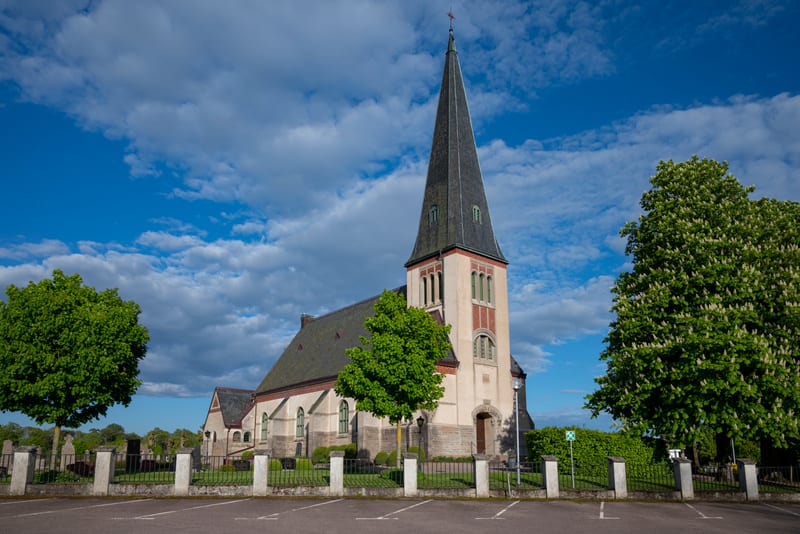 En kyrka med ett högt, smalt torn i brunt och beige under en blå himmel med vita moln. Framför kyrkan finns en asfalterad parkeringsplats med vita markeringar. Gröna träd och en kyrkogård med gravstenar syns runtomkring.