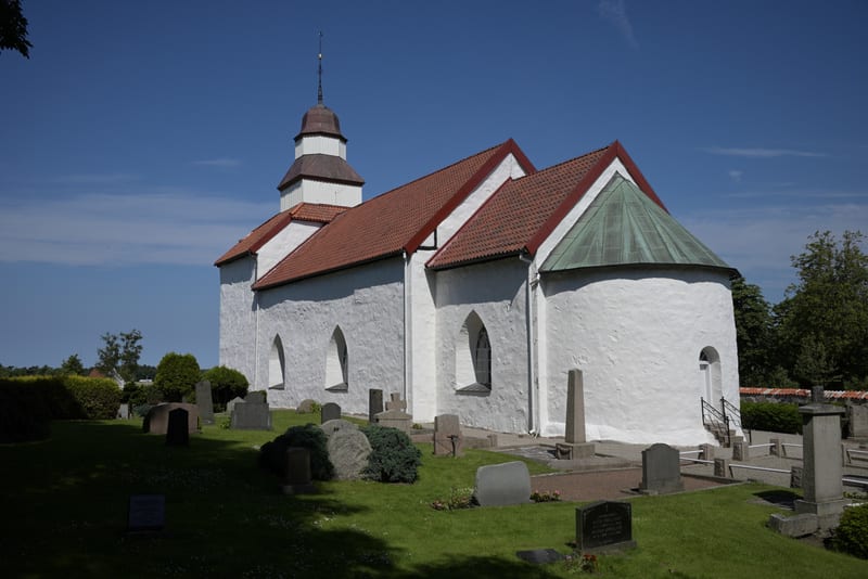 En vit kyrkbyggnad med rött tak och en tornspira syns under en molnfri blå himmel. Framför kyrkan ligger en grön gräsmatta med flera gravstenar och buskar. Byggnaden har rundade fönsteröppningar och en del av taket är grönt.