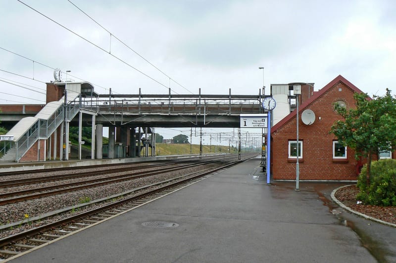 En översiktsbild av en tågstation med spår, perronger och en röd tegelbyggnad. En gångbro och trappor syns i bildens övre vänstra del. En skylt med texten ”Spår 1 Tåg mot Helsingborg och Malmö” samt en klocka är placerade vid gångbron.