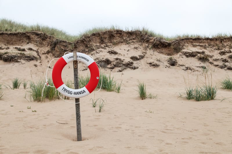 Bilden visar en ensam livboj, röd och vit med texten "LAHOLMS KOMMUN" och "TRYGG-HANSA", monterad på en stolpe i sanden. I bakgrunden syns sanddyner med gräs och en ljusgrå himmel. Små grästuftor växer även i förgrunden.
