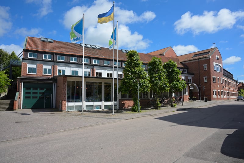 En stor, röd tegelbyggnad med tegeltak och flera fönsterrader under en blå himmel med vita moln. Framför byggnaden syns två flaggstänger med flaggor med grön och blå färg, samt en flaggstång med en gul och blå flagga. En asfalterad gata sträcker sig
