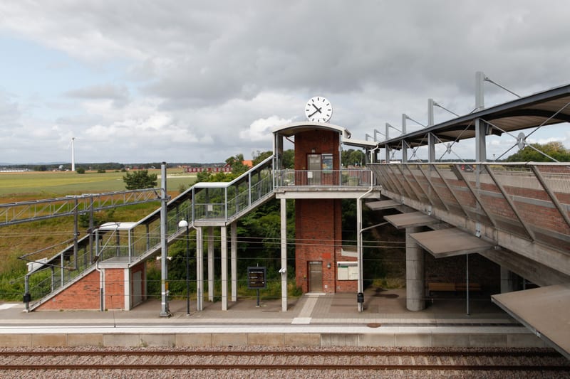 En bild som visar en järnvägsstation med ett övergångsplan med trappor och en klocka. I bakgrunden syns skog, åkrar och hus under en molnig himmel, samt ett vindkraftverk till vänster. På perrongen syns spår och stationsutrustning.