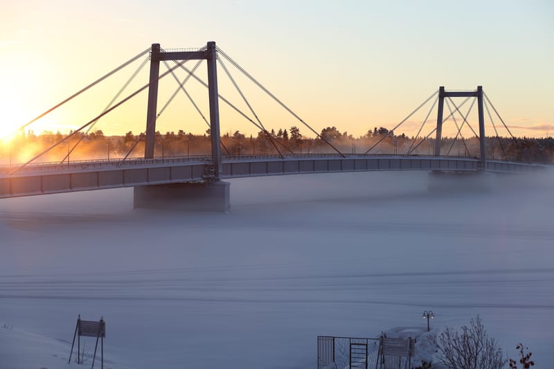 En bro i vinterlandskap med soluppgång. Dimma ligger över den snötäckta marken under bron. Himlen är gul-orange vid horisonten och ljusblå högre upp. Träd är synliga uppe på flodbanken bakom bron.