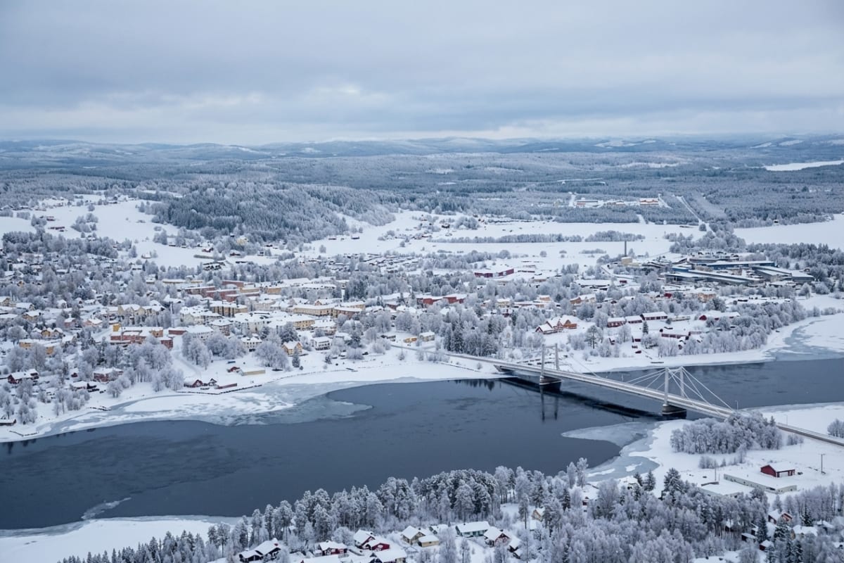 En vidvinkelbild från ovan visar ett snötäckt landskap med ett samhälle längs en delvis isig älv. En bro korsar älven, och det finns bostadshus, träd och en stor byggnad klädda i snö. Molnigt väder.