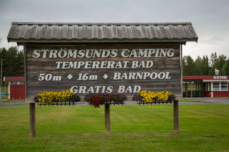 En stor, rustik träskylt med texten "STRÖMSUNDS CAMPING", "TEMPERERAT BAD", "50m", "16m", "BARNPOOL" och "GRATIS BAD". Skylten är prydd med blommande blomlådor i gult, rött och lila. I bakgrunden syns gräs, träd och en byggnad med texten "KIOSK".