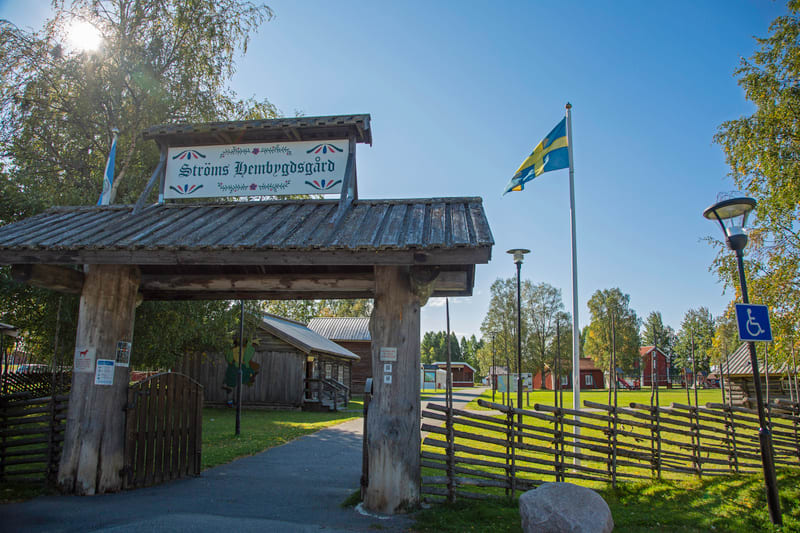 En bild visar entrén till Stöms Hembygdsgård med en skylt ovanför en trägrind. En svensk flagga vajar på en stång till höger om entrén. Ett inhägnat område med flera röda och träfärgade byggnader syns i bakgrunden under en klarblå himmel.