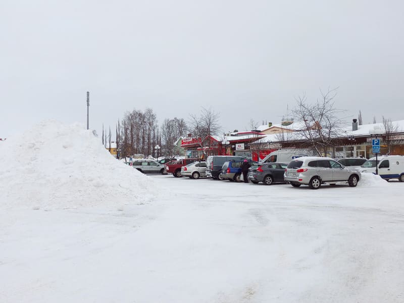 En vinterbild som visar en snötäckt parkeringsplats med flera bilar. En stor snöhög syns i förgrunden till vänster. I bakgrunden syns flera byggnader med snö på taken och trädrader med kala träd.