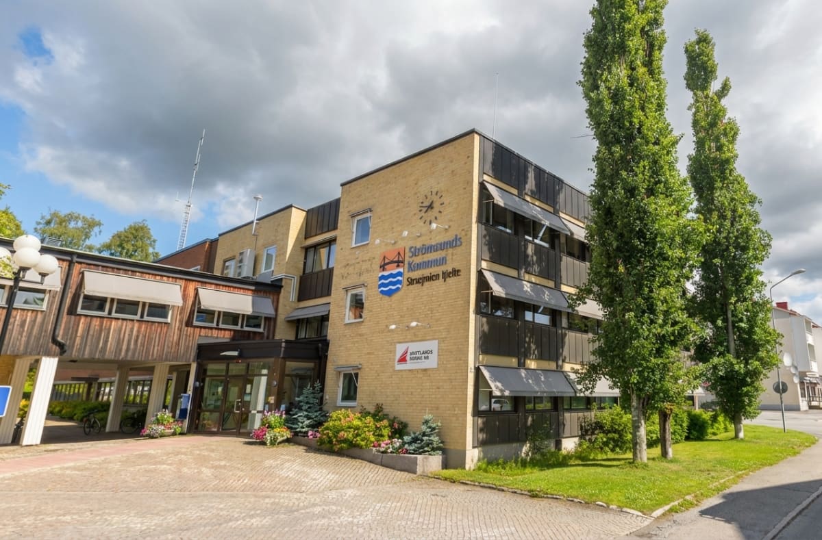 En gul tegelbyggnad med mörka detaljer och fönster med ljusa markiser, framförallt ett trähus med fönster och växter. På byggnaden syns skyltar med texten "Strömsunds Kommun" och "Jämtlands Södra AB". Träd och buskar ramar in byggnaden. En kullersten