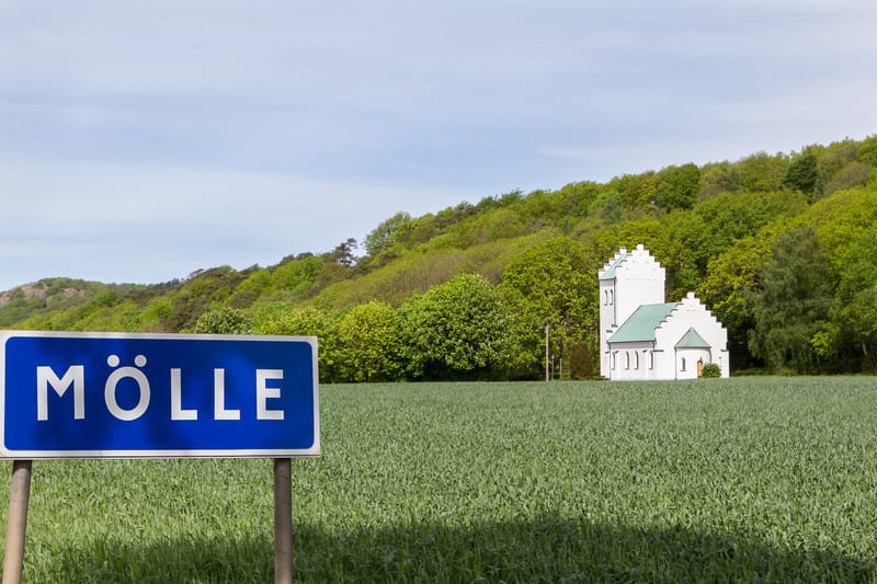 En blå skylt med texten "MÖLLE" i vitt framför ett grönt fält. I bakgrunden syns en vit byggnad med ett grönt tak, omgiven av lövträd på en sluttning under en himmel med några moln.