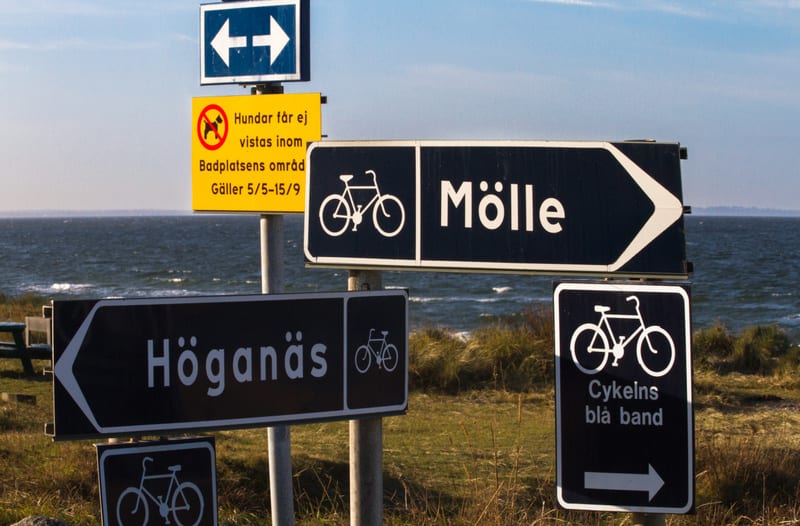 En bild som visar flera skyltar med text och symboler. I bakgrunden syns ett hav med vågor under en klarblå himmel. Marken består av gräs och buskage. En skylt förbjuder hundar på badplatsen mellan 5/5 och 15/9. Andra skyltar pekar mot Mölle och Höga