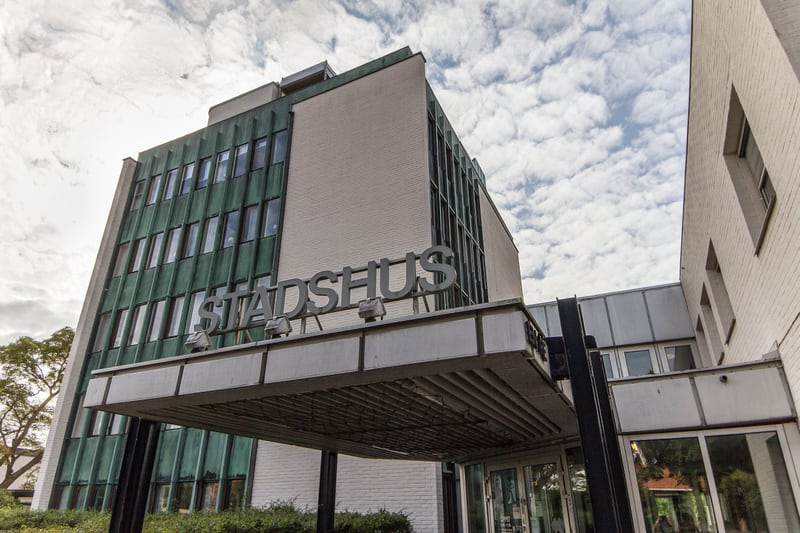 En bild som visar en del av en byggnad med texten "STADSHUS" på fasaden, sedd nedifrån och upp mot en molnig himmel. Byggnaden har vita tegelväggar och grönfärgad fasad med många fönster. Ett utskjutande tak med armaturer leder fram till entrén.