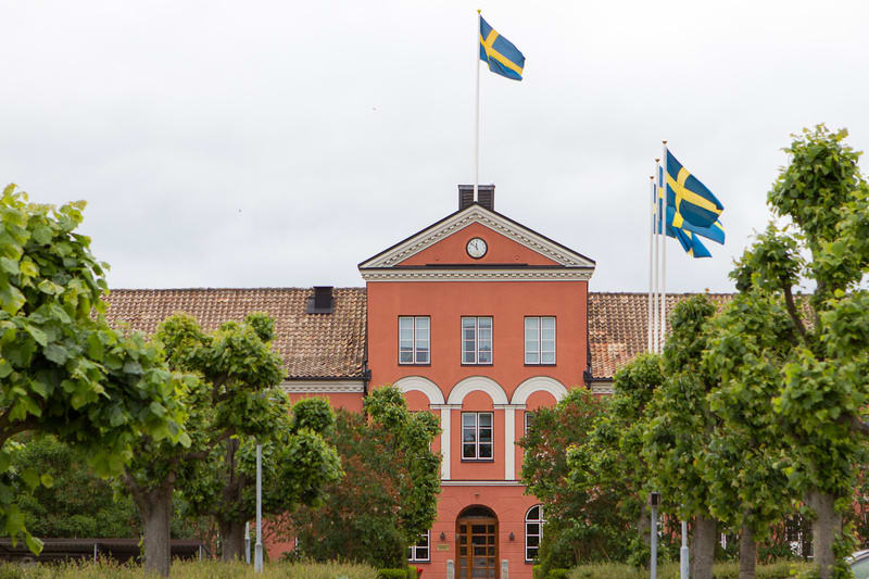 En röd byggnad med ett tegeltak under en mulen himmel. På taket syns två svenska flaggor i topp och en klocka på fasaden. Byggnaden omges av gröna träd och buskar som delvis skymmer fasaden.