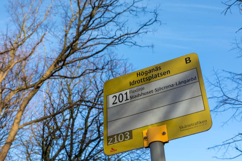 En gul busskylt syns delvis mot en klarblå himmel. Sklyten markerar "Höganäs Idrottsplatsen", "Flexlinje 201" och "Stadshuset-Sjöcrona-Långaröd". Trädgrenar utan löv är synliga i bakgrunden till vänster och i överkant. På skylten finns även Skånetraf