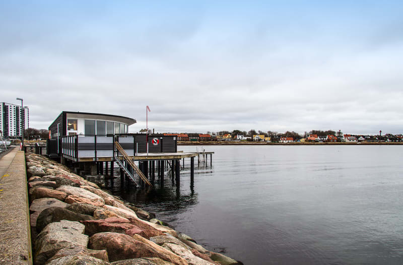 En bild som visar en byggnad på pålar vid vattnet, med en stenkant i förgrunden och ett bostadsområde på andra sidan vattnet under en molnig himmel. Byggnaden är svart och vit med stora fönster, och en röd-vit flagga syns på taket. En livboj hänger v