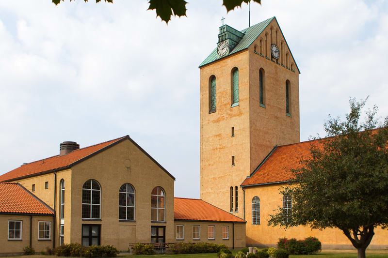 En bild som visar en kyrkobyggnad med en hög, gulaktig kyrktorn och tegelrött tak. Gröna träd och buskar syns runt byggnaden, och himlen är ljus med vita moln, samt några löv uppifrån som hänger ner.
