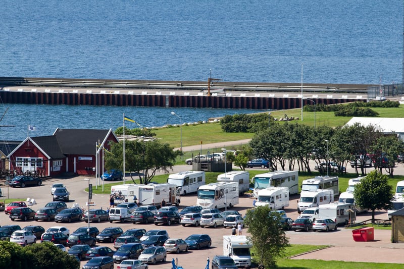 En översiktsbild av en fullsatt parkeringsplats vid en hamn, med husbilar och personbilar. Bredvid parkeringsplatsen finns en röd byggnad med svart tak och gröna ytor med träd. Längre bort syns en hamnpir och en stor vattenyta.
