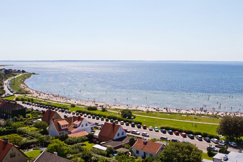 En högvinkelbild av en strandkant med många människor som badar och solar. Långs en väg med bilar parkerade och hus med röda tak. Vattnet sträcker sig till horisonten under en klarblå himmel.