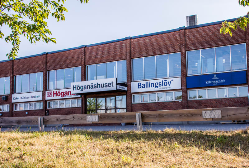 En röd tegelbyggnad under en klarblå himmel med vita skyltar på fasaden. På skyltarna läses "Höganäshuset", "Höganäs", "Ballingslöv", "Villeroy & Boch" och "Kakel • Klinker • Badrum". Framför byggnaden finns en träräcke och torrt gräs.