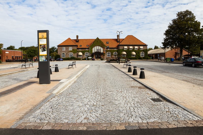 En utomhusbild som visar en stor, tegelröd byggnad med inslag av grön murgröna på fasaden. Framför byggnaden finns en öppen plats med gatsten och ett antal bänkar, samt en digital skylt. En gata med parkerade bilar syns till höger.