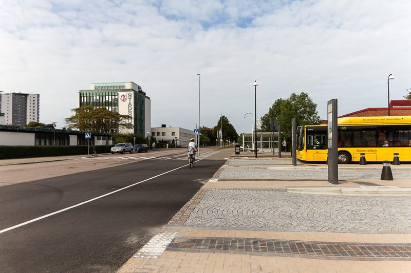En gata med en cyklist och ett buss-stopp. I bakgrunden syns flera byggnader, varav en flervåningsbyggnad med texten "STADS" på fasaden. En gul buss står vid buss-stoppet och flera personer syns i bilden. Marken består av asfalt, kullersten och beton