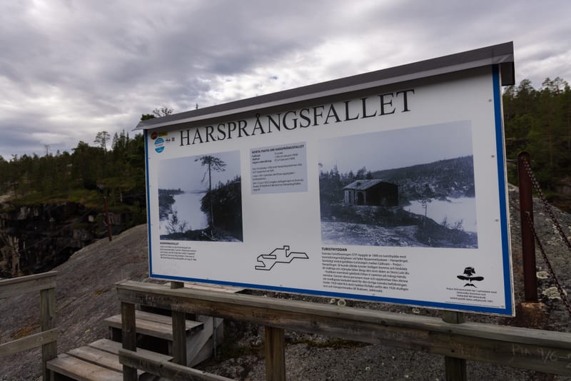 En informationsskylt om Harsprångsfallet står utomhus på en berghäll. Skylten, som har rubriken 'HARSPRÅNGSFALLET', visar svartvita bilder och text. I förgrunden syns en träkonstruktion med trappsteg, och i bakgrunden skymtar träd och en gråmulen him