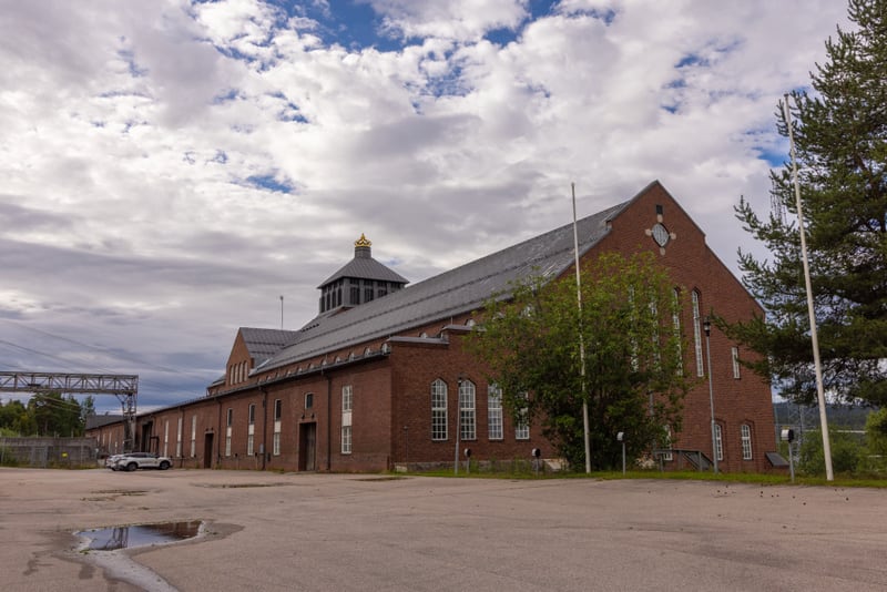En stor, röd tegelbyggnad med ett grått tak dominerar bilden under en molnig himmel. På taket syns en mörk kupol med guldfärgad topp. Framför byggnaden finns en asfalterad yta med en vattenpöl och i bakgrunden skymtar träd och industristrukturer.
