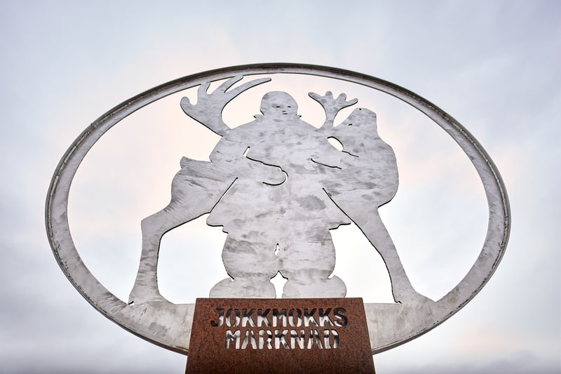En metallskulptur föreställande en mänsklig figur och två renar, placerad under en gråmulen himmel. Skulpturen är inkapslad i en oval ring och har texten "JOKKMOKKS MARKNAD" utskuren i basen av rostfärgad metall. Den mänskliga figuren bär traditionel