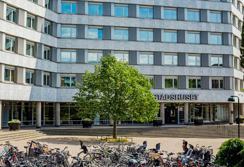 En stor, ljus byggnad med flera våningar och många fönster. Framför byggnaden finns entrén med texten "STADSHUSET" ovanför. Ett träd med gröna löv står framför entrén, och en stor samling cyklar syns i förgrunden.