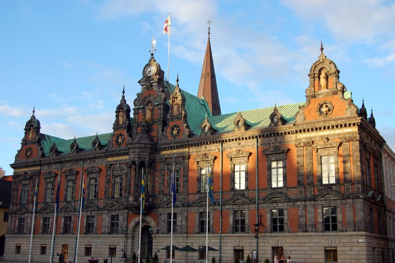 En stor, tegelröd byggnad med inslag av grå sten och ett grönt tak syns under en blå himmel. Byggnaden har många fönster, prydliga takdetaljer, en klocka, diverse statyer och flera flaggstänger med svenska och andra flaggor utanför entrén. I bakgrund