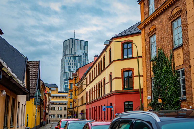 En gata kantad av flervåningshus i blandade färger, delvis skuggad. I bakgrunden reser sig en modern skyskrapa. På gatan står flera personbilar parkerade, och himlen är molnig.